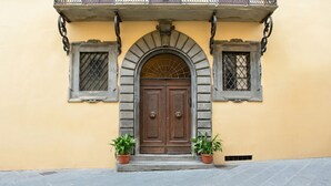 A building facade with a central arched door, flanked by two windows with metal grilles, and two potted plants at the entrance. - Loft with private bathroom (Arezzo)