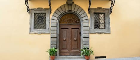 A building facade with a central arched door, flanked by two windows with metal grilles, and two potted plants at the entrance.