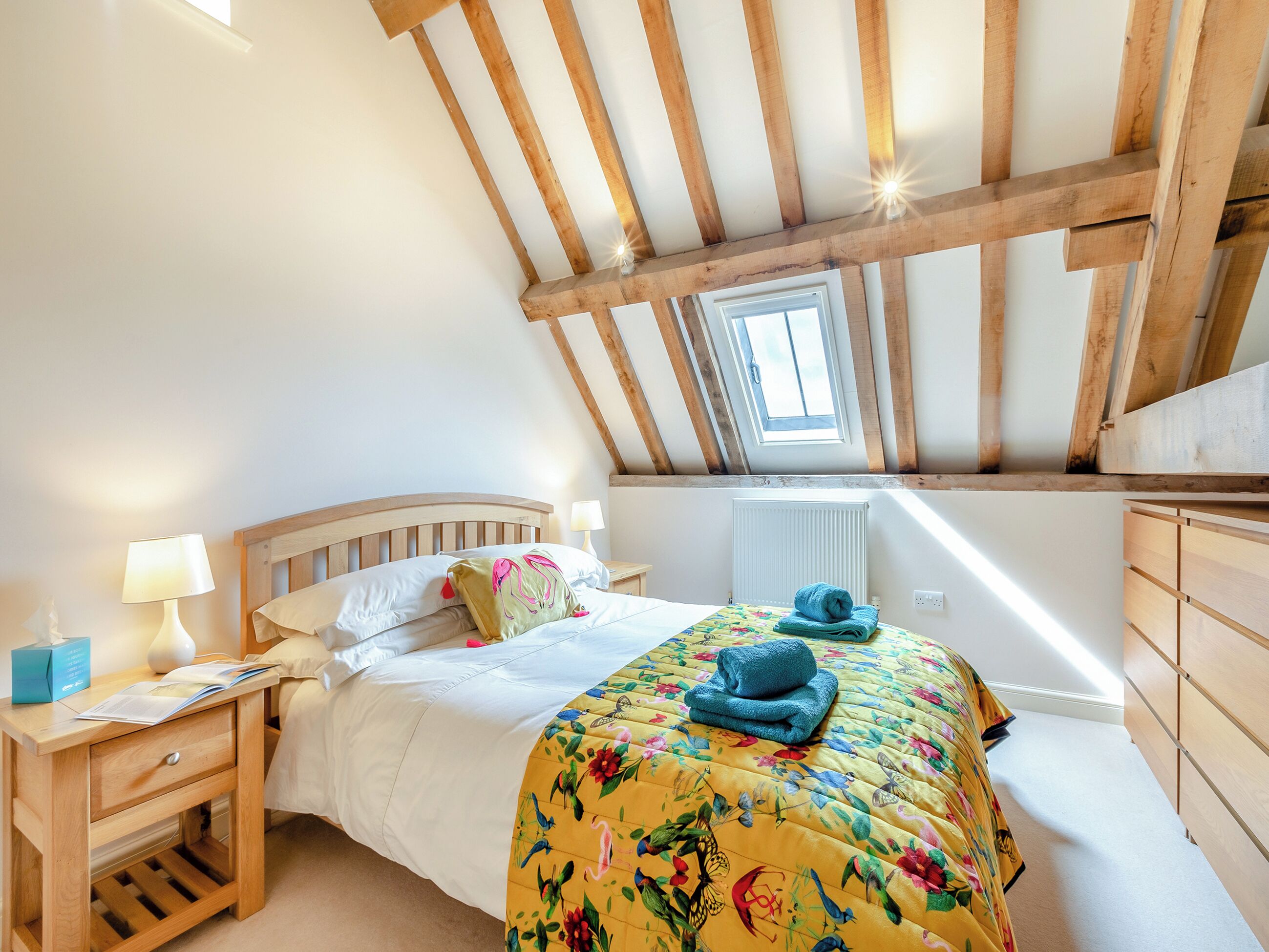 A bedroom with a sloped ceiling, a skylight, a bed with a floral bedspread, a nightstand with a lamp, and a wooden dresser.