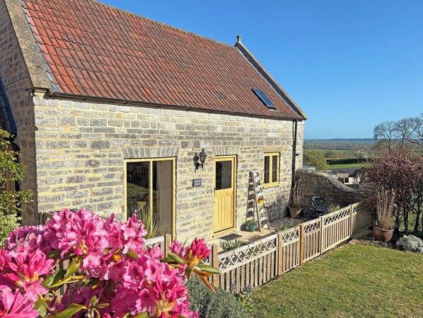 A stone house with a tiled roof, a wooden door, and a small garden with pink flowers.