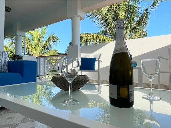 A glass table with a wine bottle and two wine glasses, a blue sofa, and a white railing with palm trees in the background.