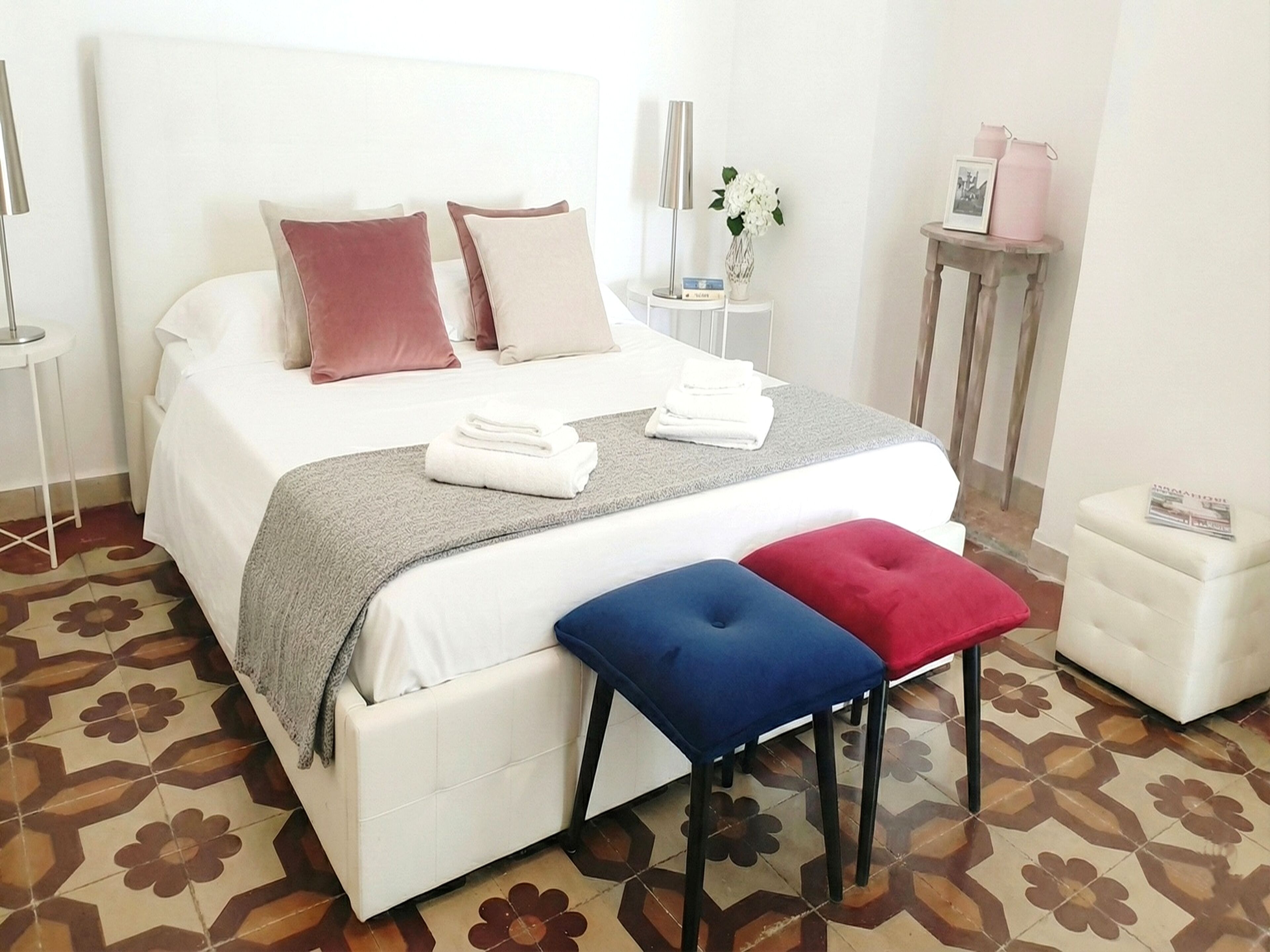 A neatly made bed with pillows, a bedside table with a lamp, a small stool, and a magazine on a side table.