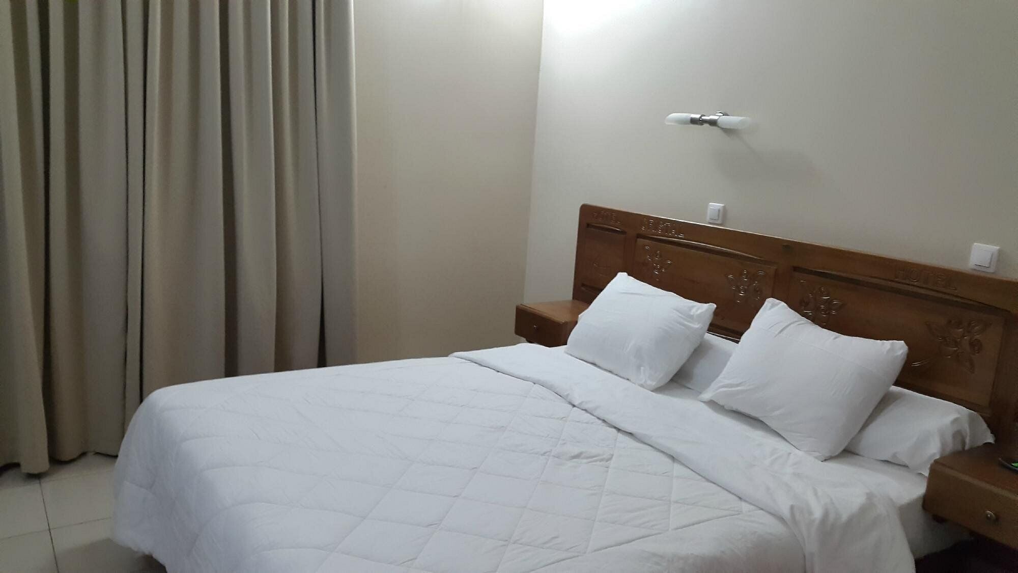 Habitación de hotel con cama, dos almohadas, mesita de noche, lámpara de pared y ventana con cortinas.
