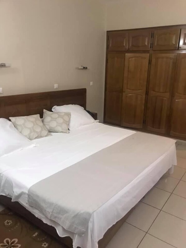 Una cama bien hecha con sábanas blancas y almohadas estampadas, un armario de madera y un piso de baldosas.