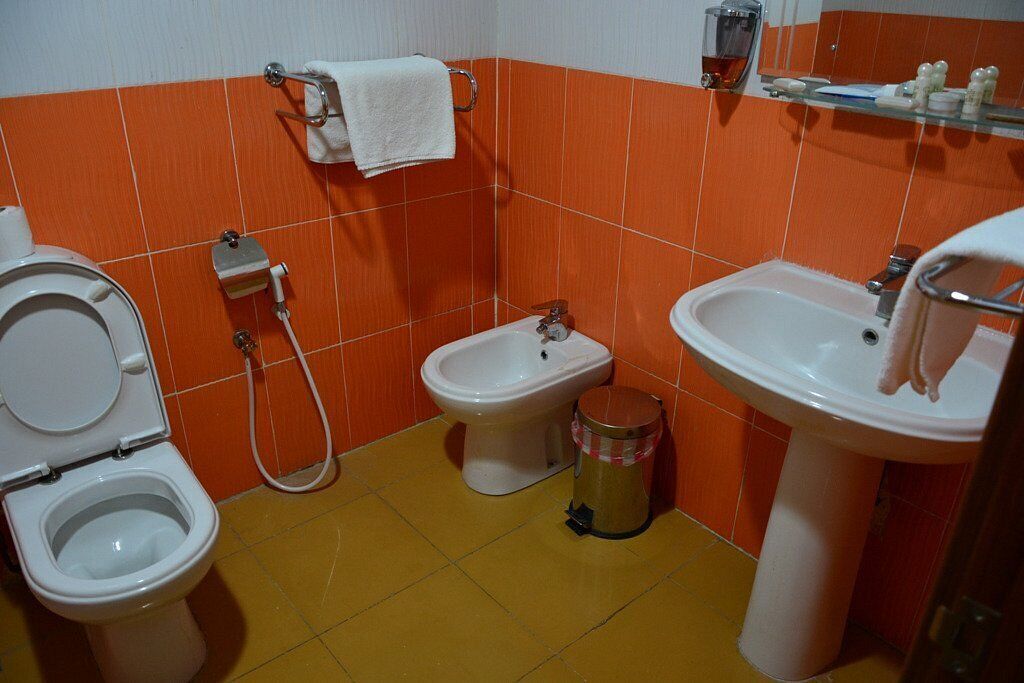 A bathroom with a toilet, bidet, and sink, orange tiled walls, and a yellow floor.