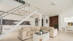 A modern living room with a beige sectional sofa, a glass coffee table, a staircase with glass railings, and a wooden door. - V196| Villa Brisas - Four Bedroom Villa, Sleeps 8 (Torre Soli Nou)