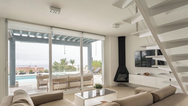 A modern living room with a sofa, a coffee table, a TV, a fireplace, and a balcony with a pool. - V196| Villa Brisas - Four Bedroom Villa, Sleeps 8 (Torre Soli Nou)