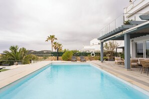 A swimming pool with lounge chairs, a covered patio, and a view of a white building with a balcony.