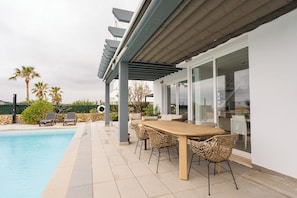 A poolside area with a wooden table and wicker chairs, a pergola, and a view of the pool.