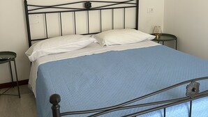 Ein Bett mit einer blauen Decke, weißen Kissen und einem Metallgestell. Es gibt einen kleinen Nachttisch mit einer Lampe und einem grünen Hocker.