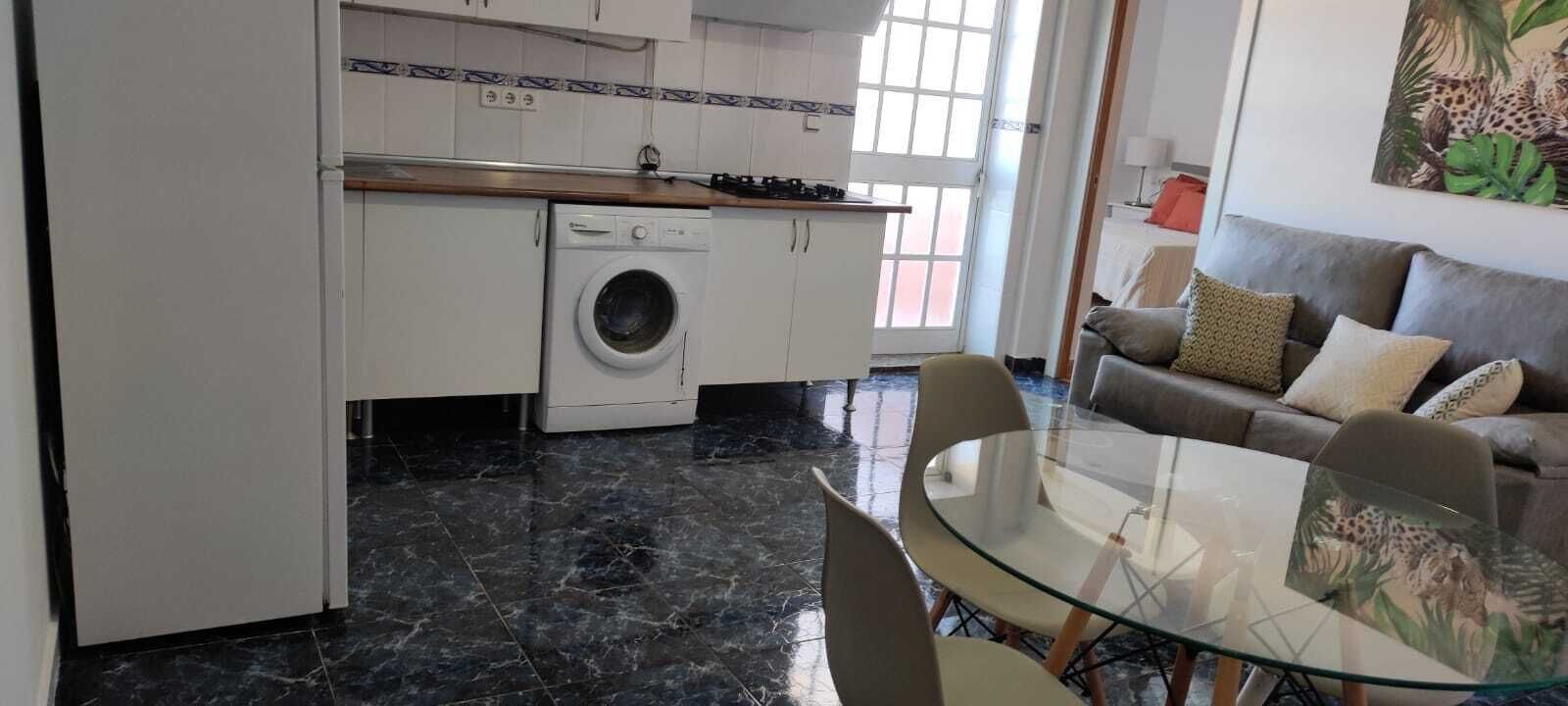 Ett kompakt kök med tvättmaskin, en matplats med ett glasbord och en soffa med kuddar.