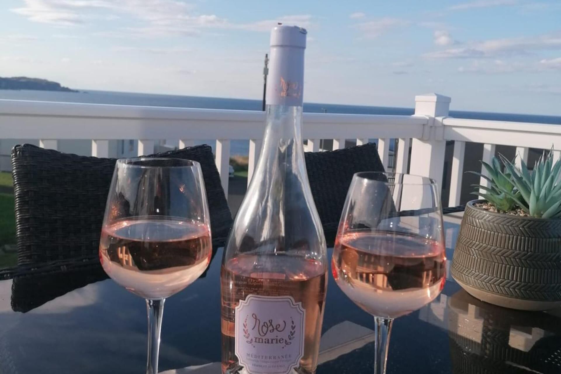 A table with two wine glasses and a bottle of rosé wine on a balcony with a view of the ocean.