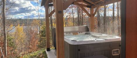 A hot tub under a wooden gazebo with a view of a lake and autumn trees.