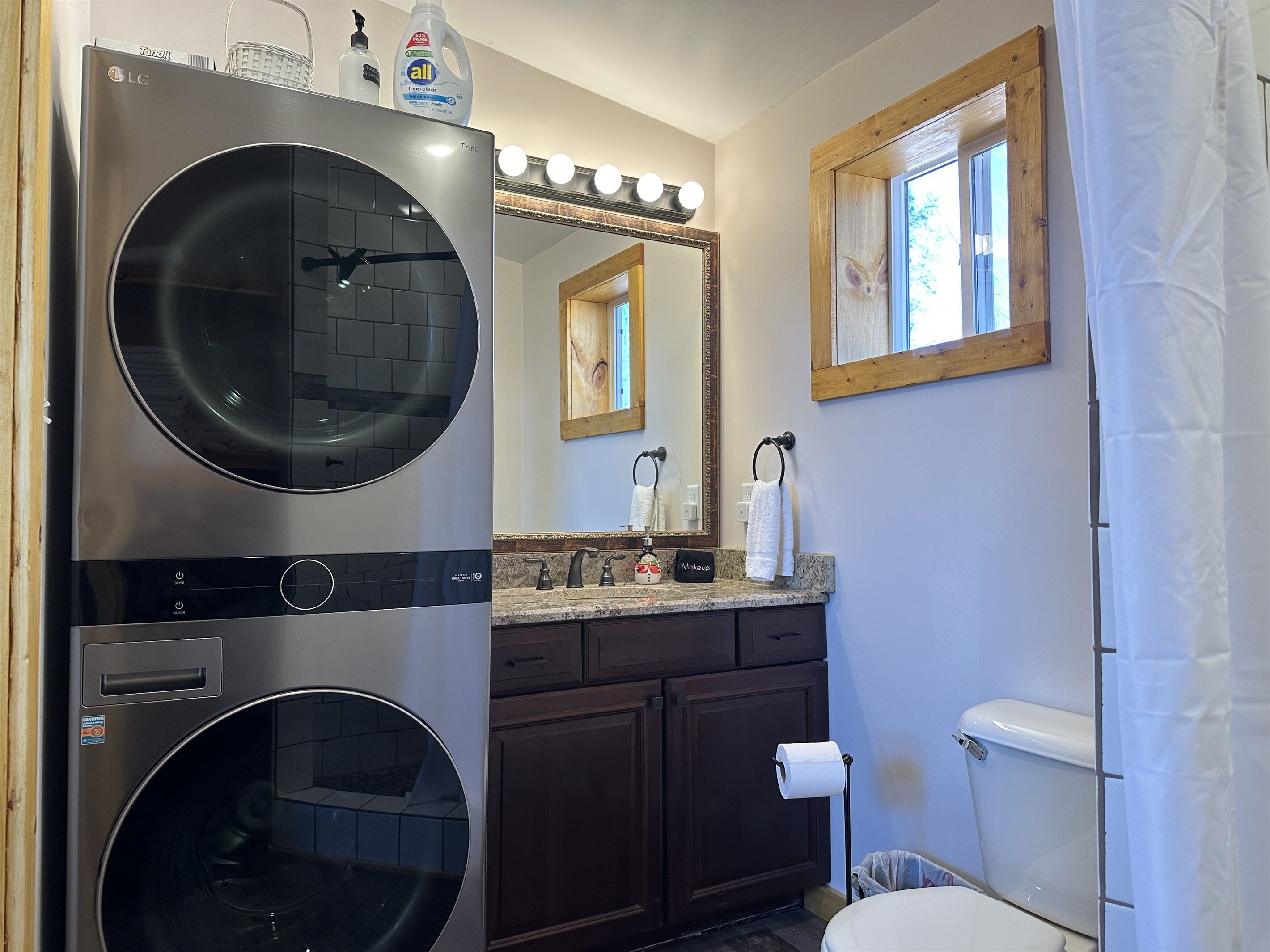 A modern bathroom with a double washer-dryer unit, a vanity with a mirror, a toilet, and a window with a wooden frame.