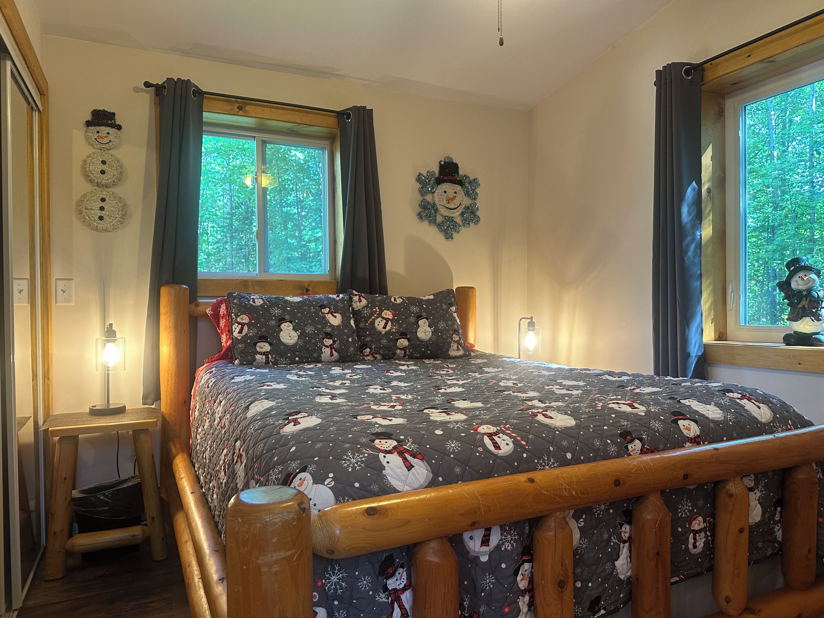 Quarto com cama estampada com tema de boneco de neve, cabeceira de madeira, duas janelas com cortinas, relógio na parede e mesa de cabeceira com abajur.