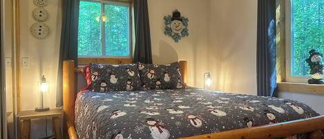 Quarto com cama estampada com tema de boneco de neve, cabeceira de madeira, duas janelas com cortinas, relógio na parede e mesa de cabeceira com abajur.