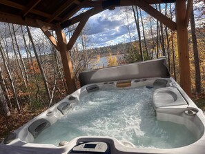 Un bain à remous doté d’un panneau de commande, entouré d’une terrasse en bois et bordé d’une zone boisée aux couleurs automnales.