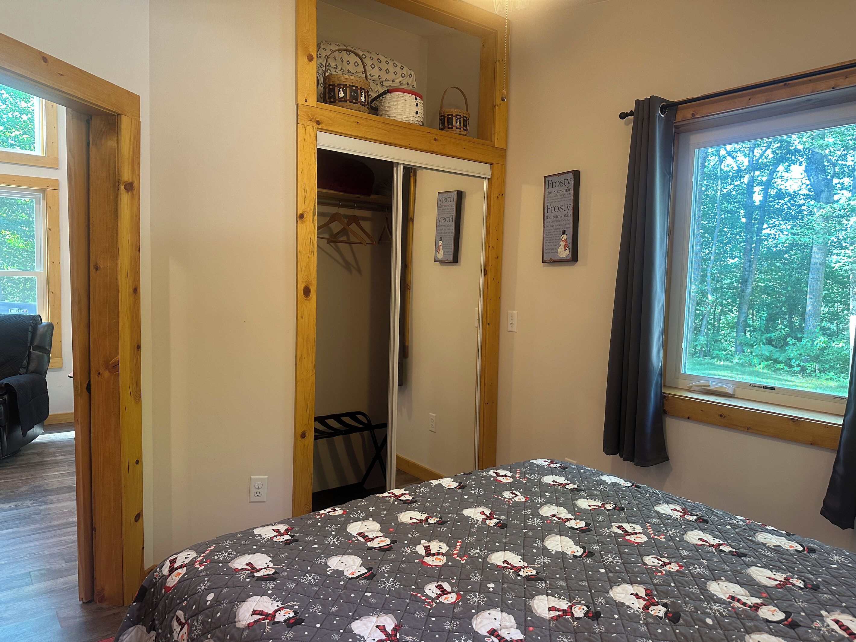Un dormitorio con una cama estampada con un diseño de muñeco de nieve, un armario de madera con ropa colgada y una ventana que ofrece vistas a un bosque.