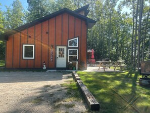 A wooden cabin with a white door and windows, a welcome sign, a snowman decoration, and a picnic table in a forest setting. - Hot tub - Great views - close to PRNL - private (Christmas)