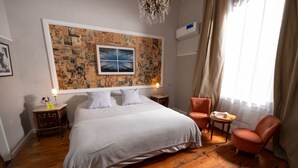 Un dormitorio con una cama grande, una silla, una mesita, una pared con un collage y una ventana con cortinas.