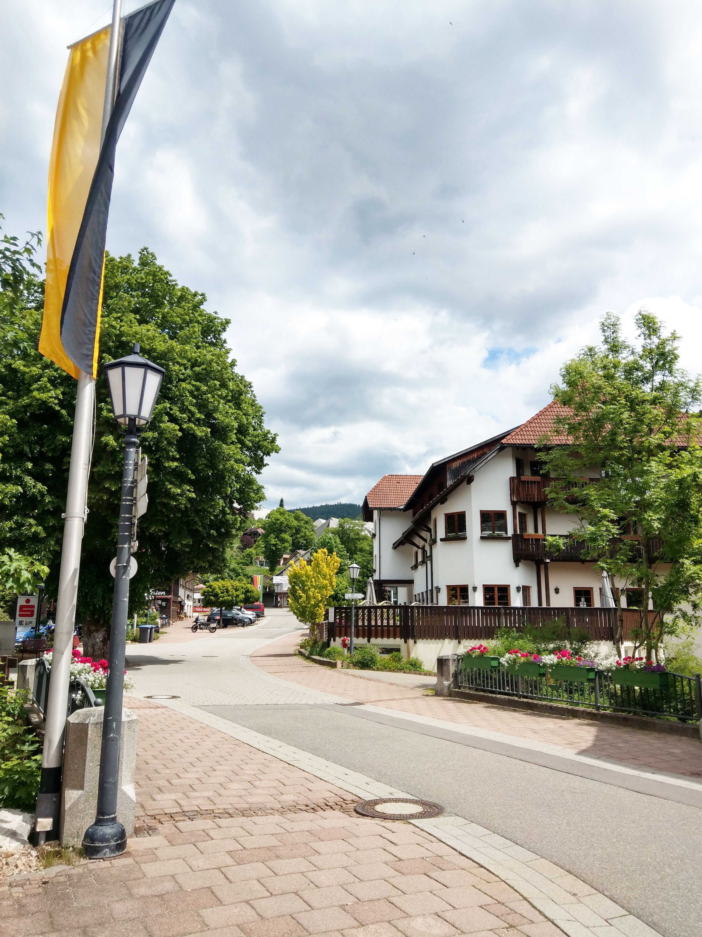 A street with a white building with a red roof, a flagpole with a yellow flag, and a lamppost.