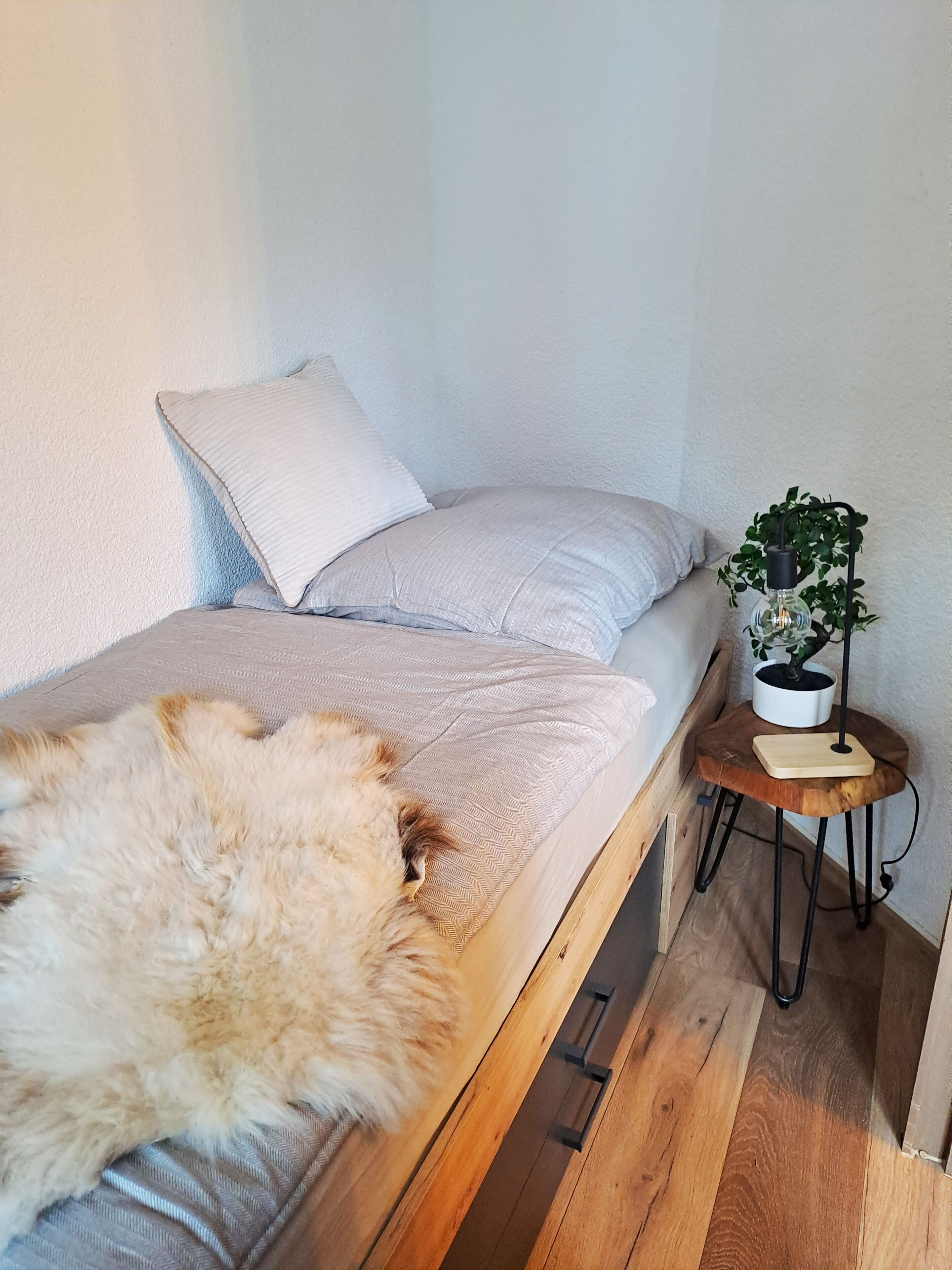Ein kleiner, gemütlicher Raum mit Bett, einer hölzernen Bank, einem Tisch mit Pflanze und einem Fellüberwurf.