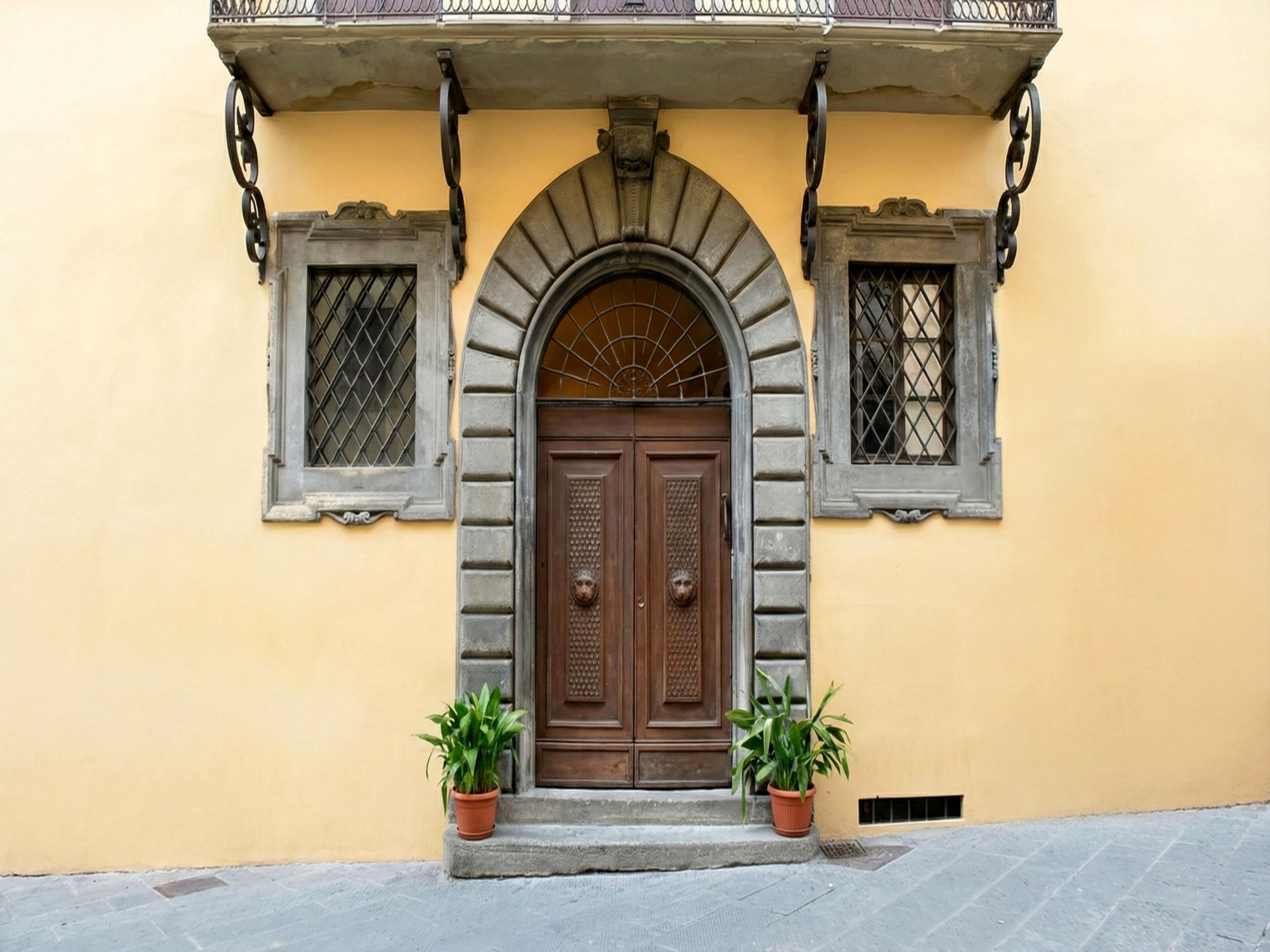 A building with a central arched entrance, flanked by two windows with metal grilles, and two potted plants on either side.
