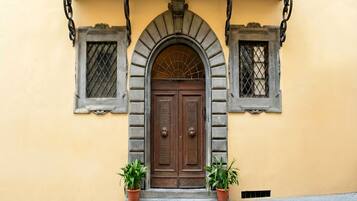 A building with a central arched entrance, flanked by two windows with metal grilles, and two potted plants on either side.