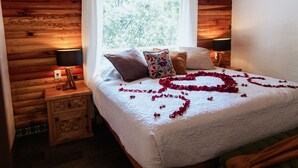 Een slaapkamer in een houten blokhut met een bed, kussens, nachtlampjes en een raam met gordijnen.
