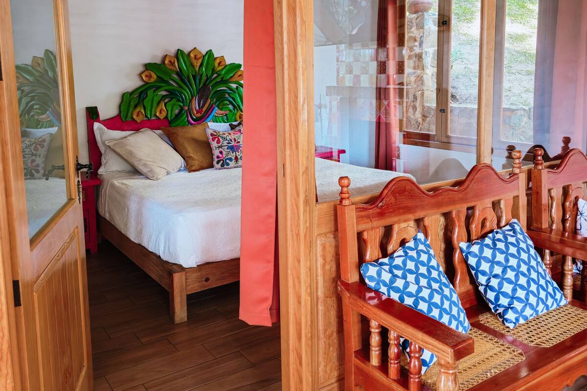 Een kamer met houten frame, een bed, een houten bank met gestreepte kussens en een grote spiegel.