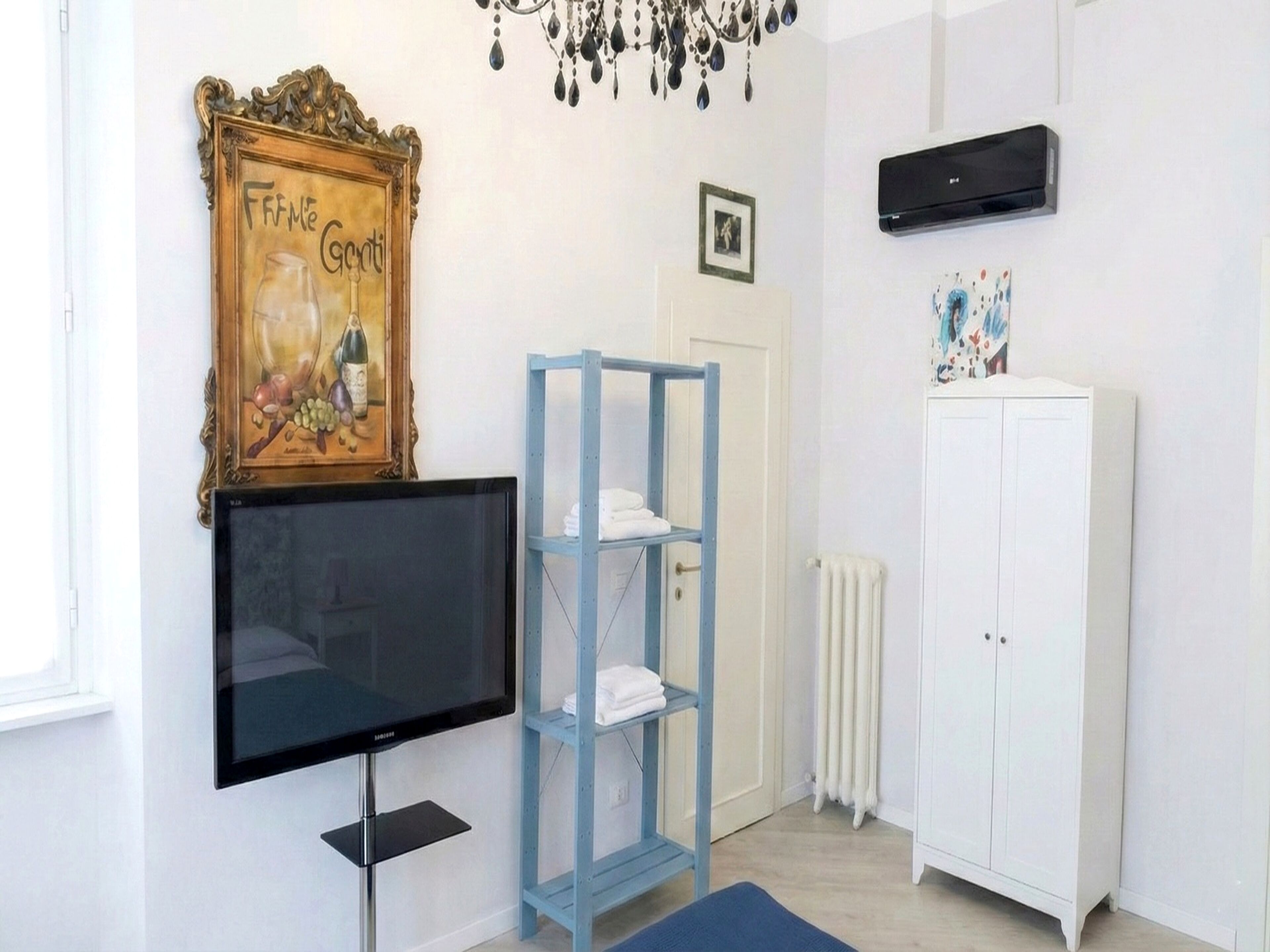 A room with a television, a framed artwork, a blue shelving unit, a white wardrobe, and a radiator.
