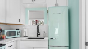 A kitchen with white cabinets, a mint green refrigerator, a vintage-style microwave, and a window with blinds. - Sunset Bungalows Lil' Green: Heated Pool, Near Beach & DT, Fenced (Clearwater)