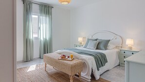 Ein Schlafzimmer mit Bett, Nachttischen, einer Pendelleuchte, einem Korbstuhl und einem Fenster mit Vorhängen.