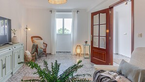 Ein geräumiges Wohnzimmer mit Fernseher, Korbmöbeln, einer großen Holztür und Blick durch ein Fenster.