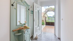 Ein Flur mit einem hellgrünem Waschtisch, einem Spiegel und einem kleinen Tisch mit einer Blumenvase. Eine Tür führt zu einem Balkon mit Blick auf einen Garten und einen Zaun.