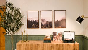 Une table en bois, avec des livres, un tourne-disque et une lampe. Trois tableaux encadrés sont accrochés au mur au-dessus.
