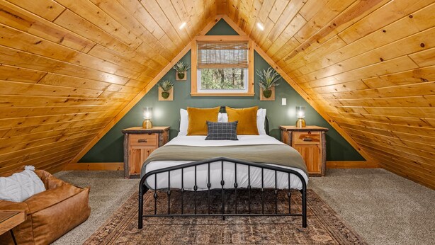 Une chambre mansardée chaleureuse, avec un plafond en bois incliné, un grand lit avec une tête de lit, deux tables de chevet avec des lampes et une vue sur la verdure par la fenêtre.