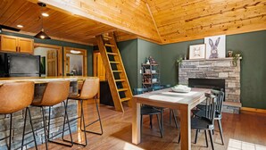 L’intérieur chaleureux d’un chalet avec un plafond en bois, une cheminée en pierre, un coin repas avec une table et des chaises, et une cuisine équipée d’un réfrigérateur et de tabourets de bar.