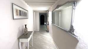 Um corredor com piso branco, um quadro emoldurado à esquerda, uma pequena mesa com um vaso e um espelho grande à direita.