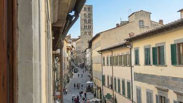 A narrow street with buildings on either side, a prominent tower in the background, and people walking.