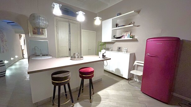 Uma cozinha com uma ilha central, dois banquinhos, uma geladeira rosa e um armário branco.
