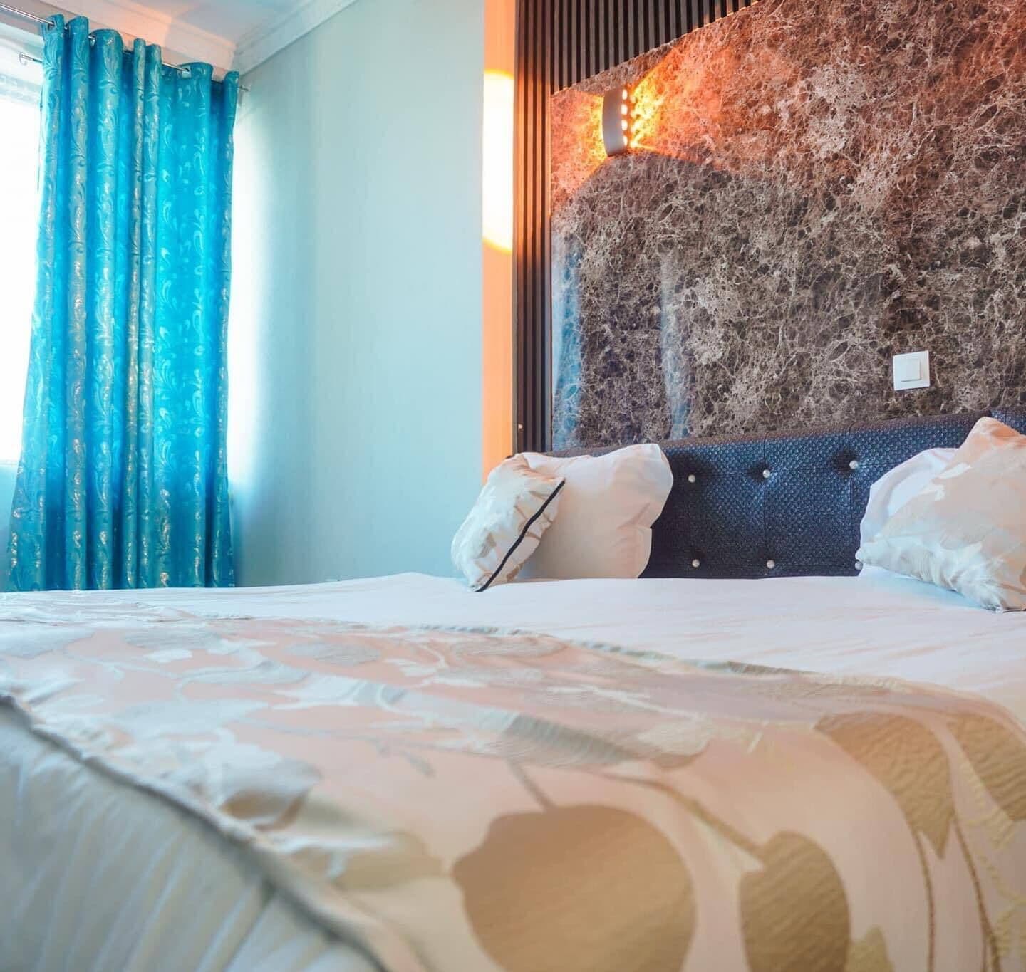 Ein Hotelzimmer mit Bett, blauen Vorhängen, einer strukturierten Wand und einem Lichtschalter.