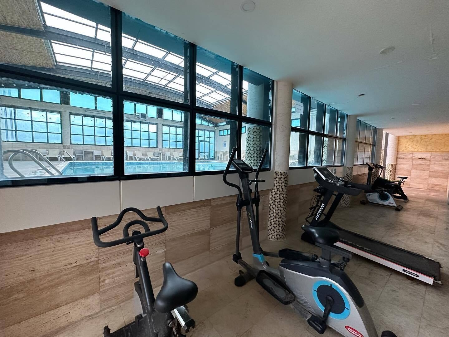 Un gimnasio con equipamiento de ejercicio, ventanas amplias y vista a una piscina.