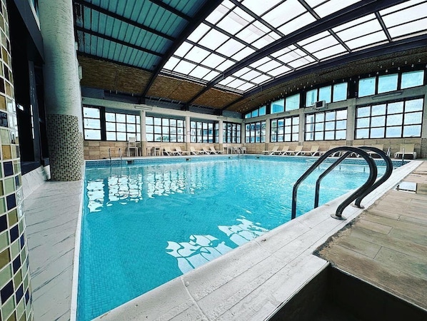 Una piscina interior con techos de vidrio transparente, paredes de azulejos y sillones de descanso alrededor.
