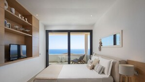 Un dormitorio moderno con una cama grande, un televisor, un balcón con vistas al mar y una estantería de madera con objetos decorativos.