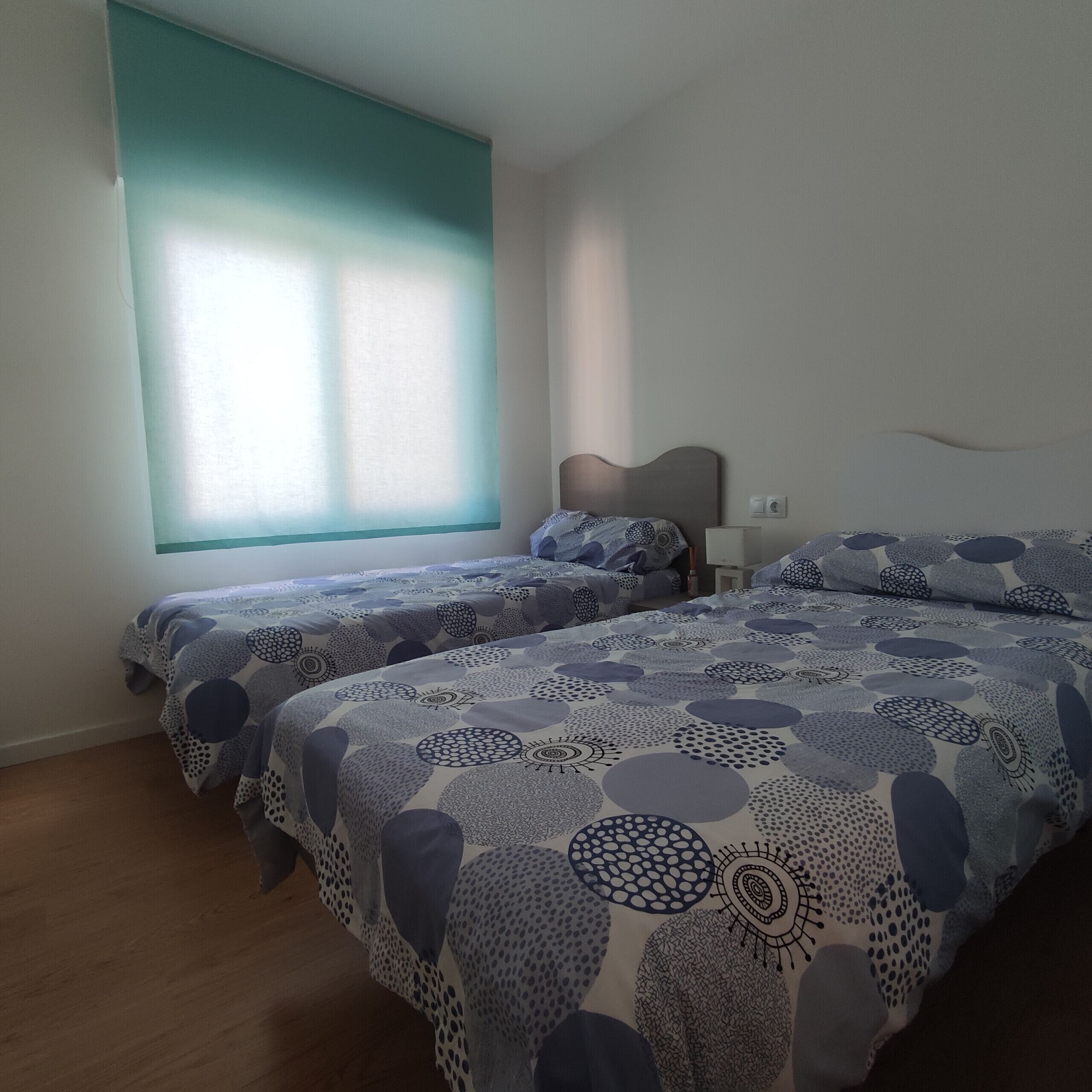 A hotel room with two beds, a window with blinds, a bedside table, and a wall socket.