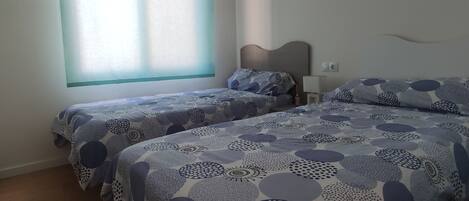 A hotel room with two beds, a window with blinds, a bedside table, and a wall socket.