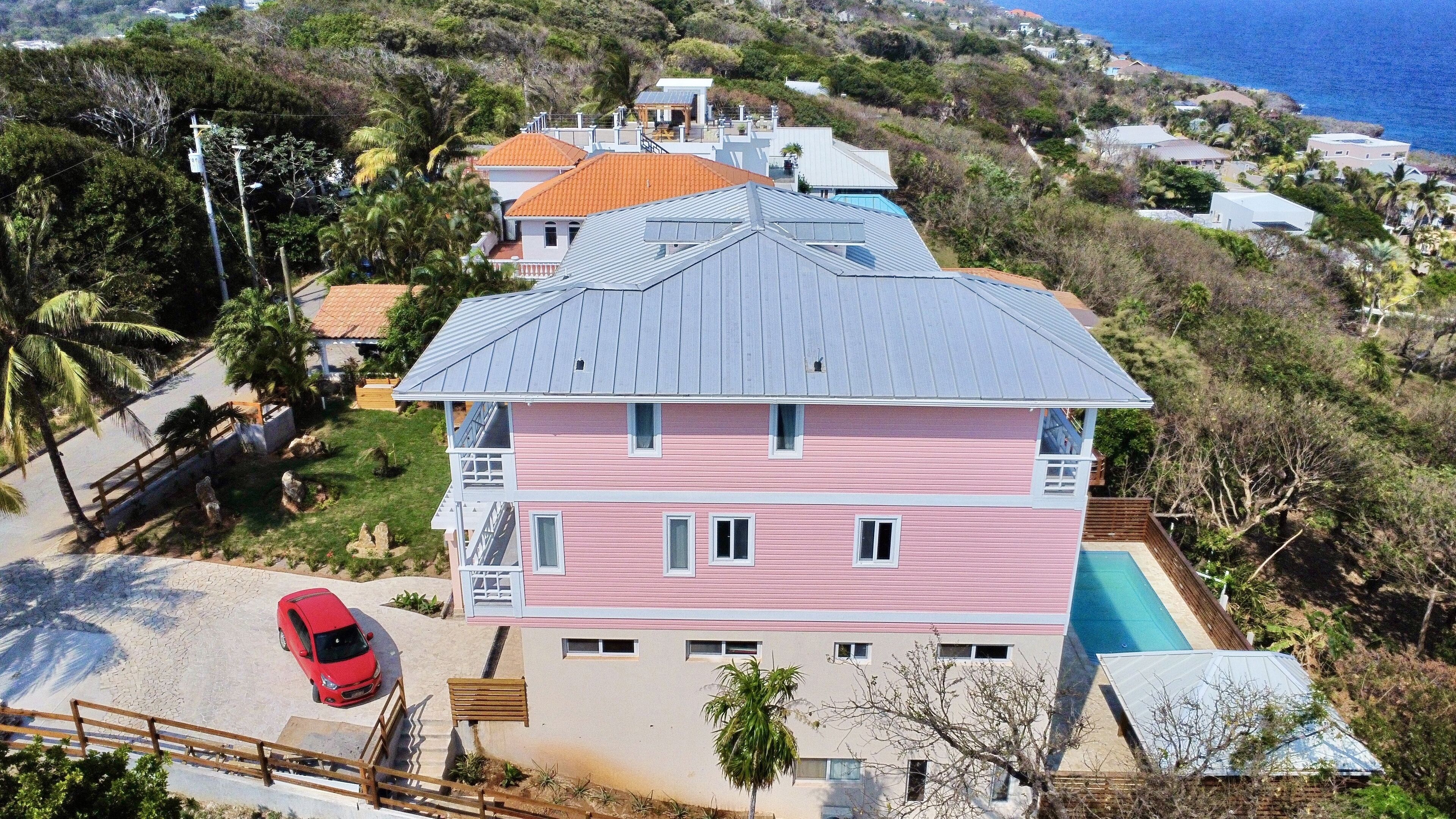 Una casa rosa de varios pisos con piscina, rodeada de exuberante vegetación y vista al océano.