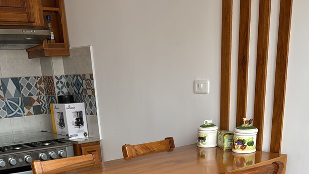 A wooden dining table with two chairs, a kitchen with a stove, and patterned floor tiles.