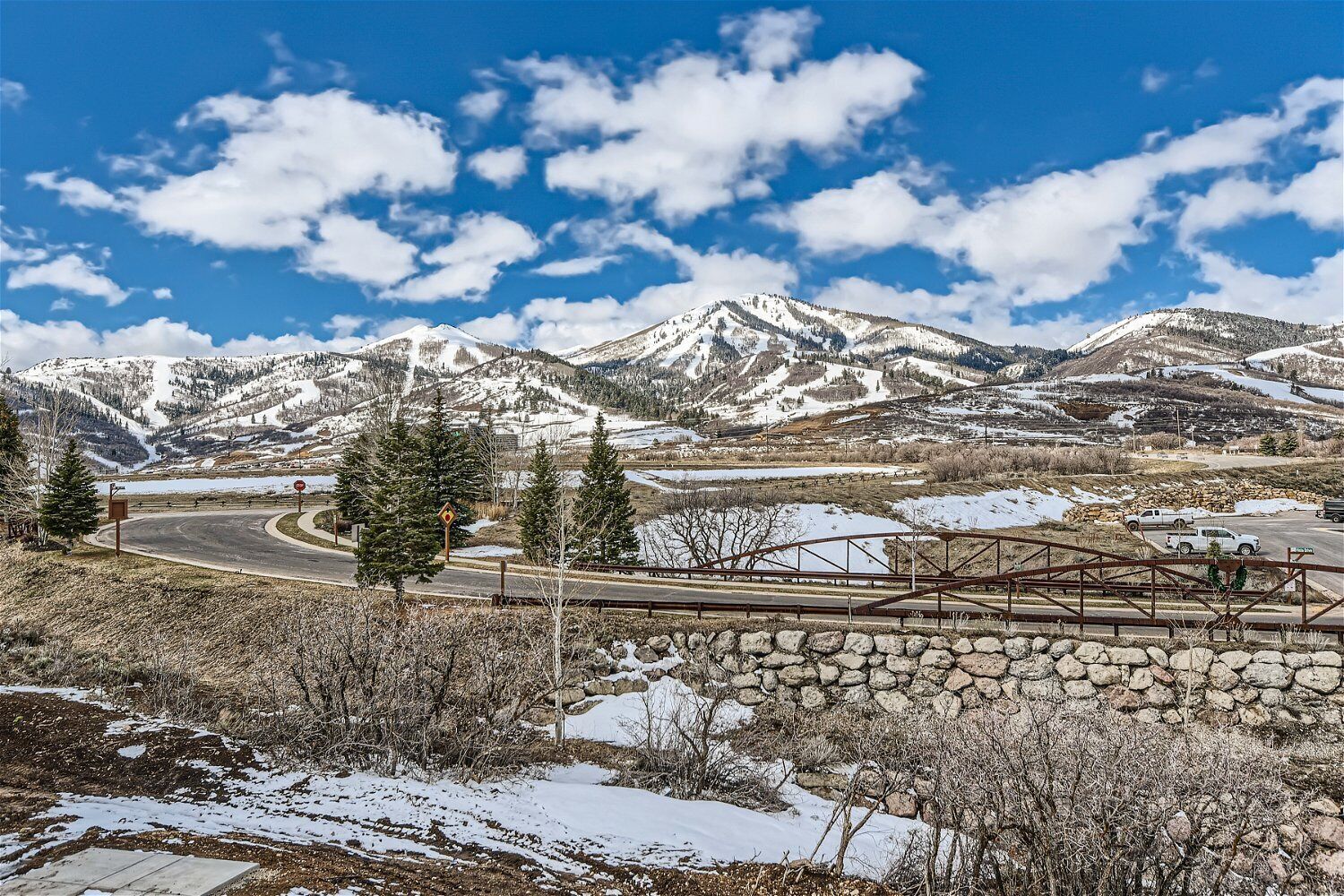 Paisaje nevado de montañas con una carretera, un muro de piedra y un puente de metal.